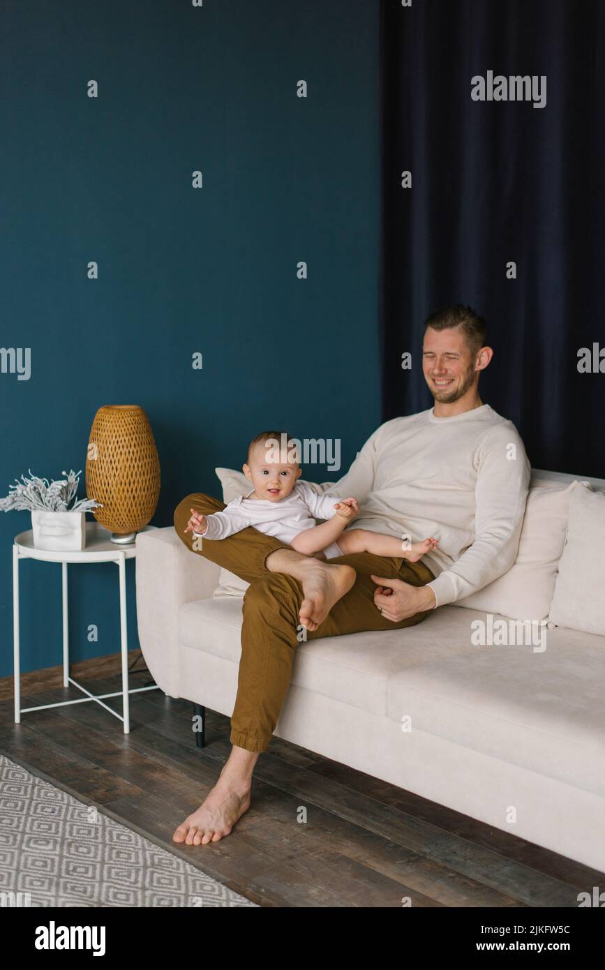 El joven es feliz y sostiene a su hijo de seis meses en sus brazos, sentado en el sofá de la habitación Foto de stock