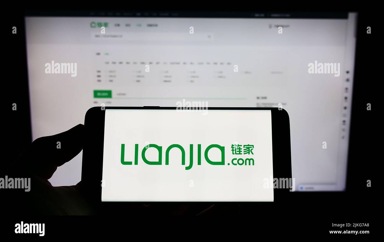 Persona que sostiene el teléfono celular con el logotipo de la compañía inmobiliaria china Lianjia en la pantalla delante de la página web del negocio. Enfoque la pantalla del teléfono. Foto de stock