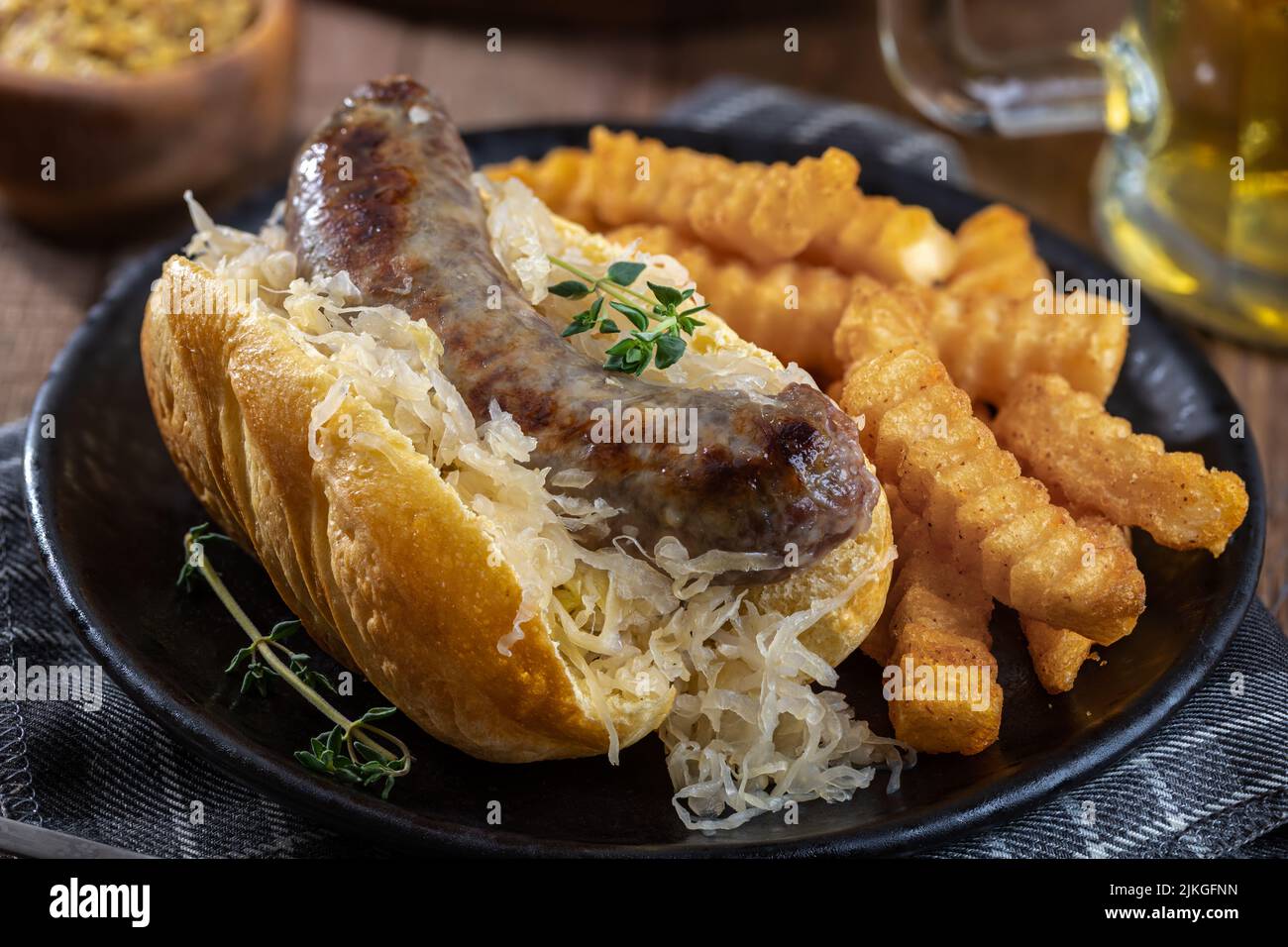 Primer plano de bratwurst a la parrilla y chucrut en un bollo con patatas fritas en un plato negro Foto de stock