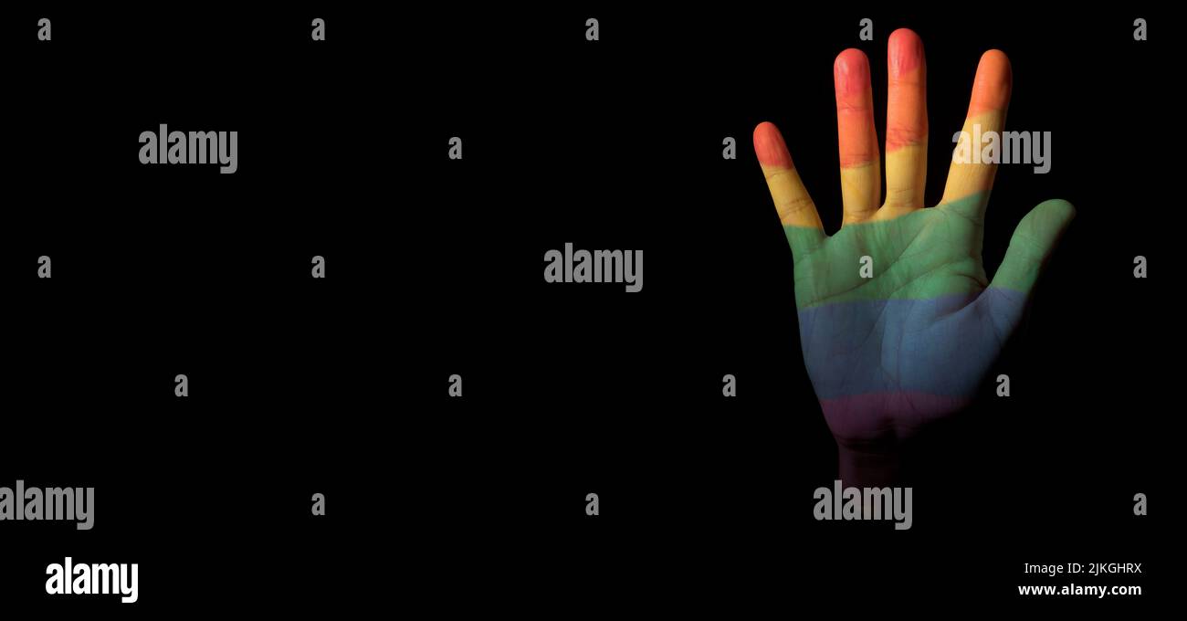 la mano de una persona, estampada con la bandera de orgullo arco iris, emerge del fondo negro, con un espacio en blanco a la izquierda, en forma panorámica Foto de stock