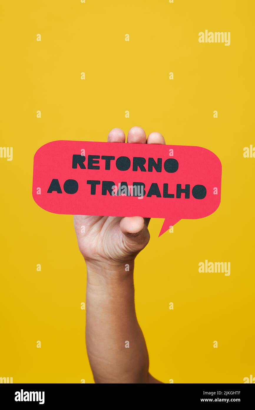 primer plano de un hombre sosteniendo una burbuja de discurso de papel rojo con el texto de nuevo a trabajar escrito en portugués, sobre un fondo amarillo Foto de stock