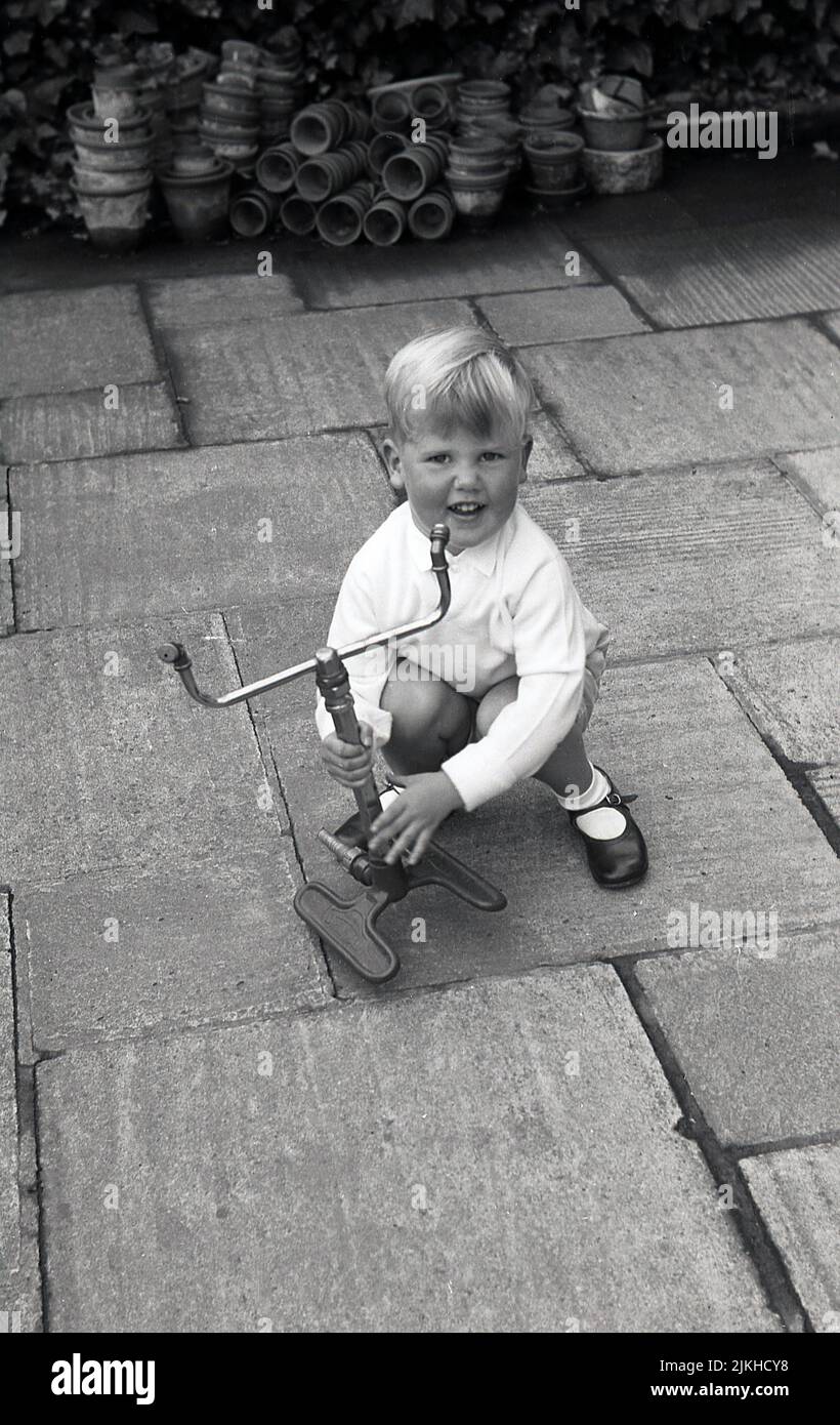 1960s, fuera en un patio, un niño pequeño feliz sosteniendo un rociador del césped del jardín de la época, hecho por Dron-Wal, de hierro fundido y latón, Inglaterra. REINO UNIDO. Foto de stock