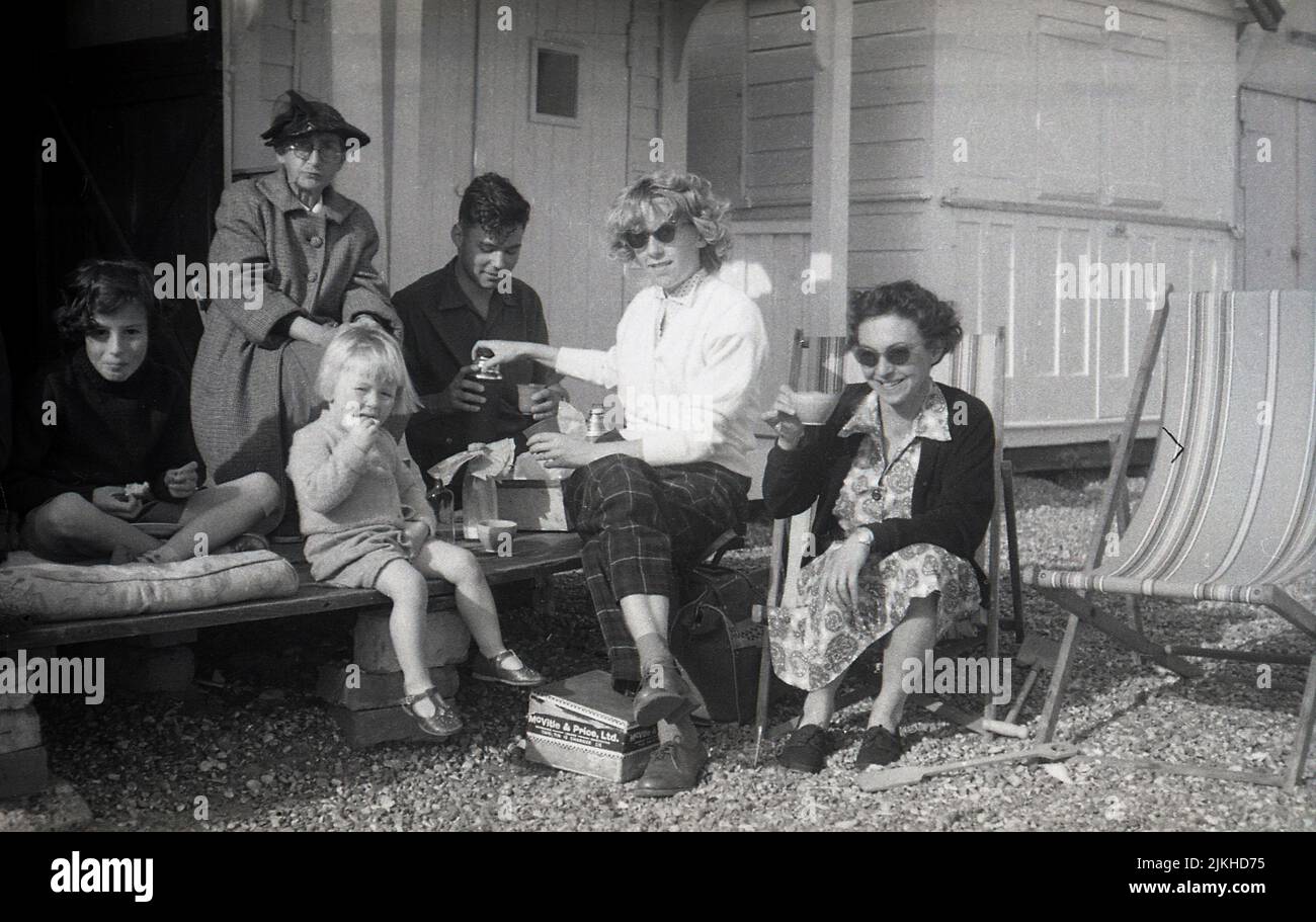 1958, histórica, una familia se reunió en la plataforma exterior de madera de su cabaña de playa, Inglaterra, Reino Unido, tomando té y sándwiches. Una caja de hojalata con la etiqueta The Mcvitie & Price Ltd se puede ver en los guijarros. Foto de stock