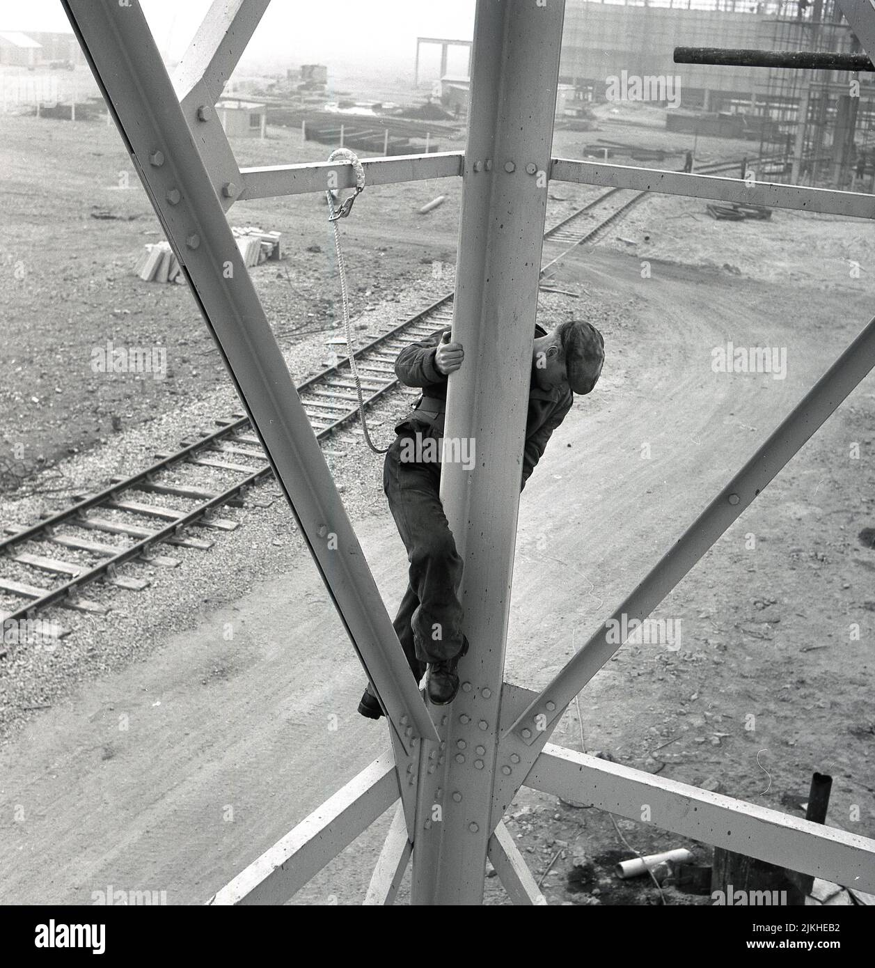 1950s, histórico, un trabajador de una gran acería, en lo alto del suelo, subiendo en el exterior de una estructura de acero enmarcado, con una cuerda de seguridad atada a él, tapa de tela en la cabeza, Abbey Works, Port Talbot, Gales. REINO UNIDO. Foto de stock
