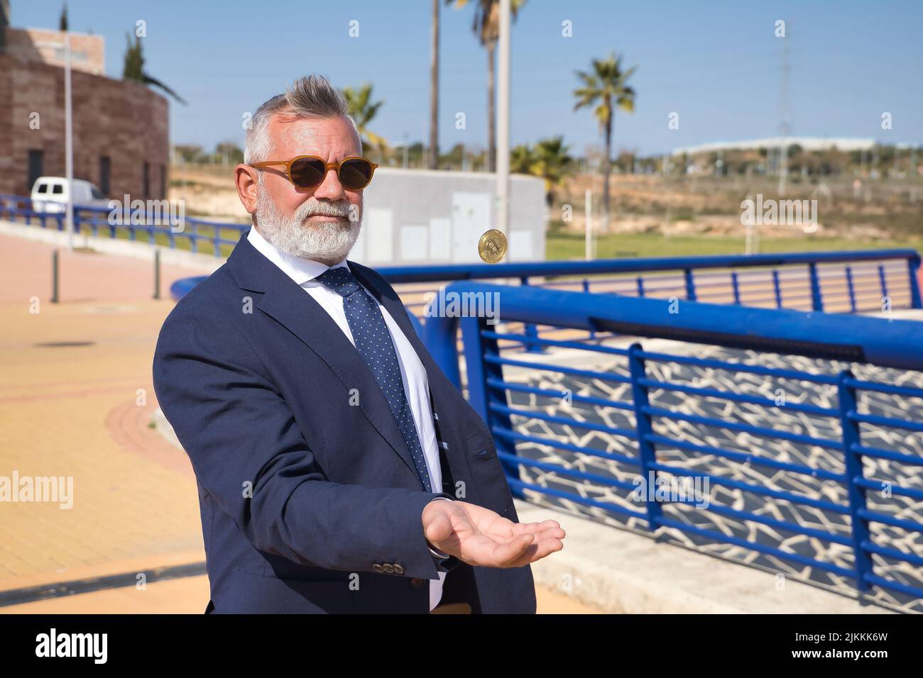 Hombre maduro, ejecutivo, de pelo gris, barbudo, con gafas de sol, chaqueta y corbata, lanzando una criptomoneda en el aire con la mano. Concepto ejecutivo Foto de stock