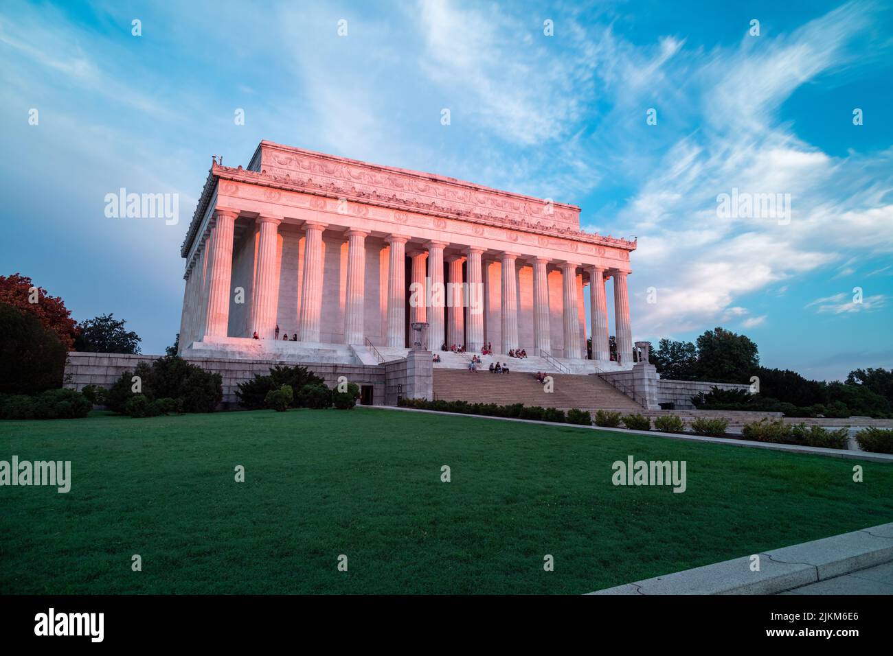La luz rosa del amanecer ilumina las columnas orientadas al este y el exterior del Lincoln Memorial en Washington, DC. En el escalón se sientan varias personas que hacen turismo. Foto de stock