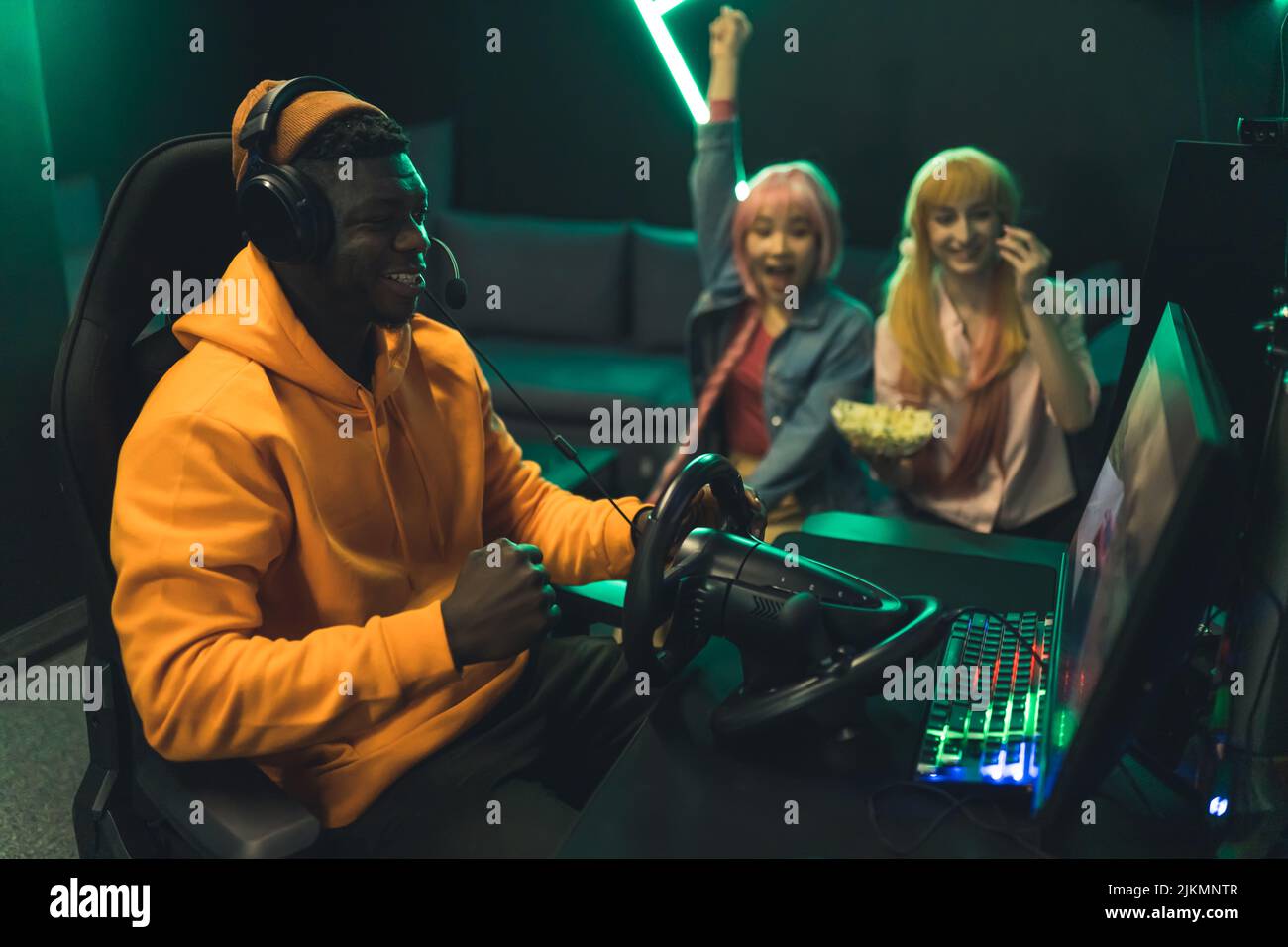 Adolescentes celebrando ganar un juego de carreras. Adolescente negro con capucha naranja y auriculares sentado en una silla de juegos. Configuración profesional con volante. Fotografía de alta calidad Foto de stock