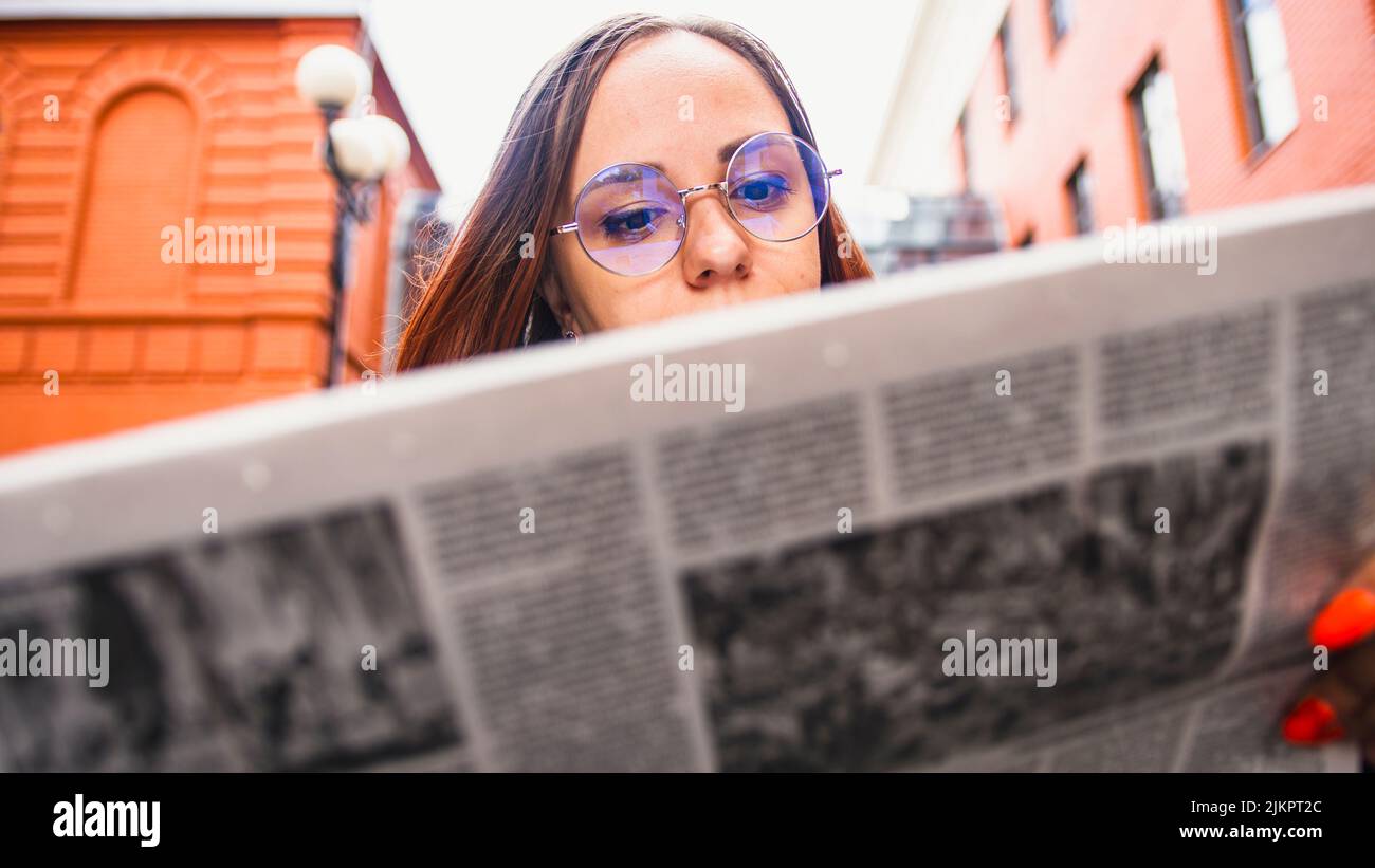 Joven pensativa leyendo el periódico en la calle. Mujer joven empresaria confundida con pelo largo marrón en ropa formal y gafas de pie Foto de stock
