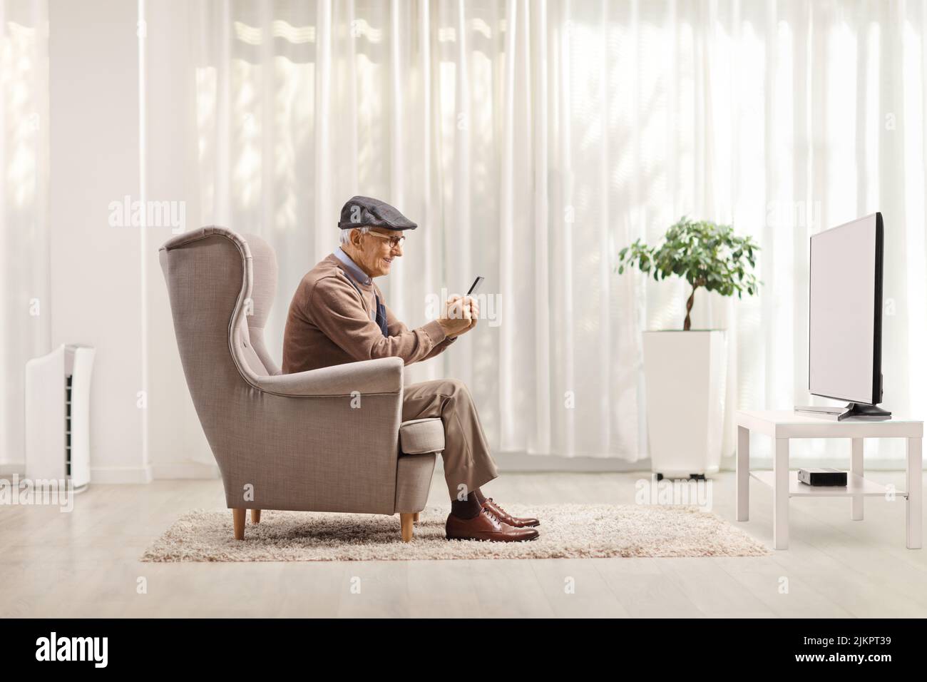 Hombre mayor en un sillón sentado frente a un televisor y usando un smartphone en casa Foto de stock