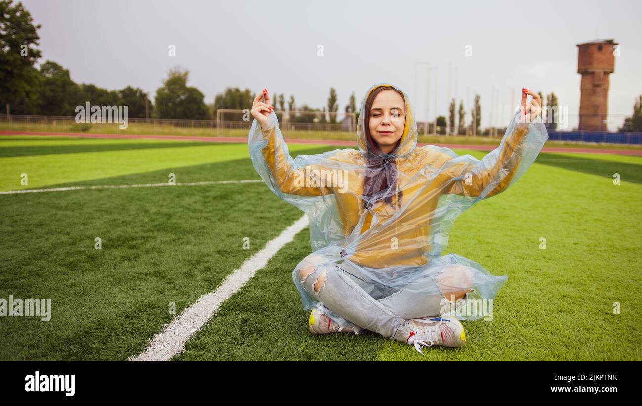 Cuerpo completo de mujer joven feliz con pelo largo marrón en ropa casual y impermeable transparente de plástico sentado en verde césped verde hierba y meditando wi Foto de stock
