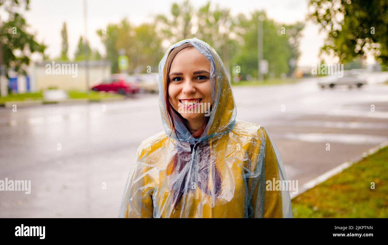 Mujer joven encantada sonriendo a la cámara el día lluvioso. Alegre mujer joven milenaria con capucha amarilla y impermeable transparente al borde de la carretera Foto de stock