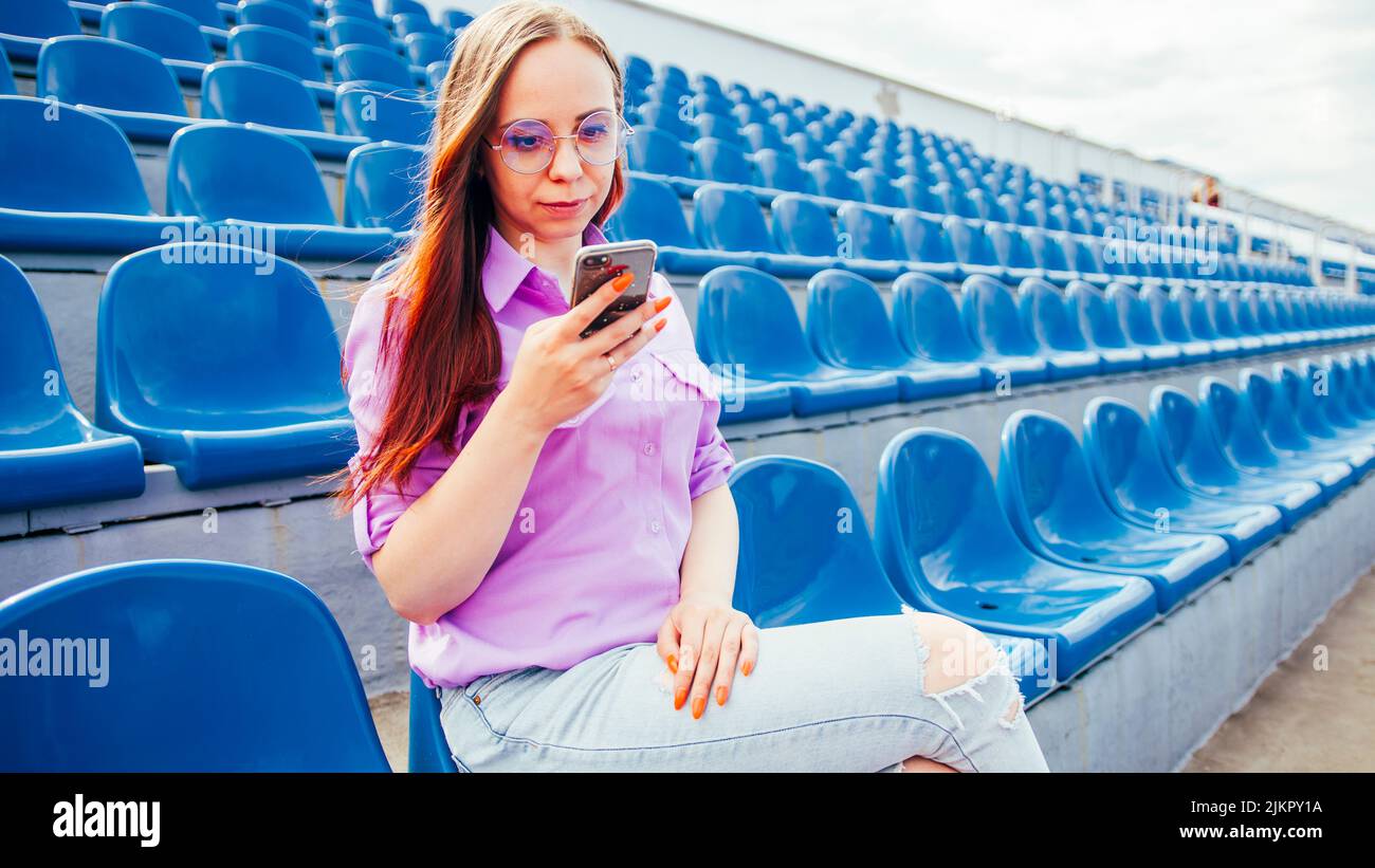 Mujer adulta concentrada con pelo largo marrón en blusa y gafas sentado en el asiento de la tribuna azul y mensajería en smartphone Foto de stock