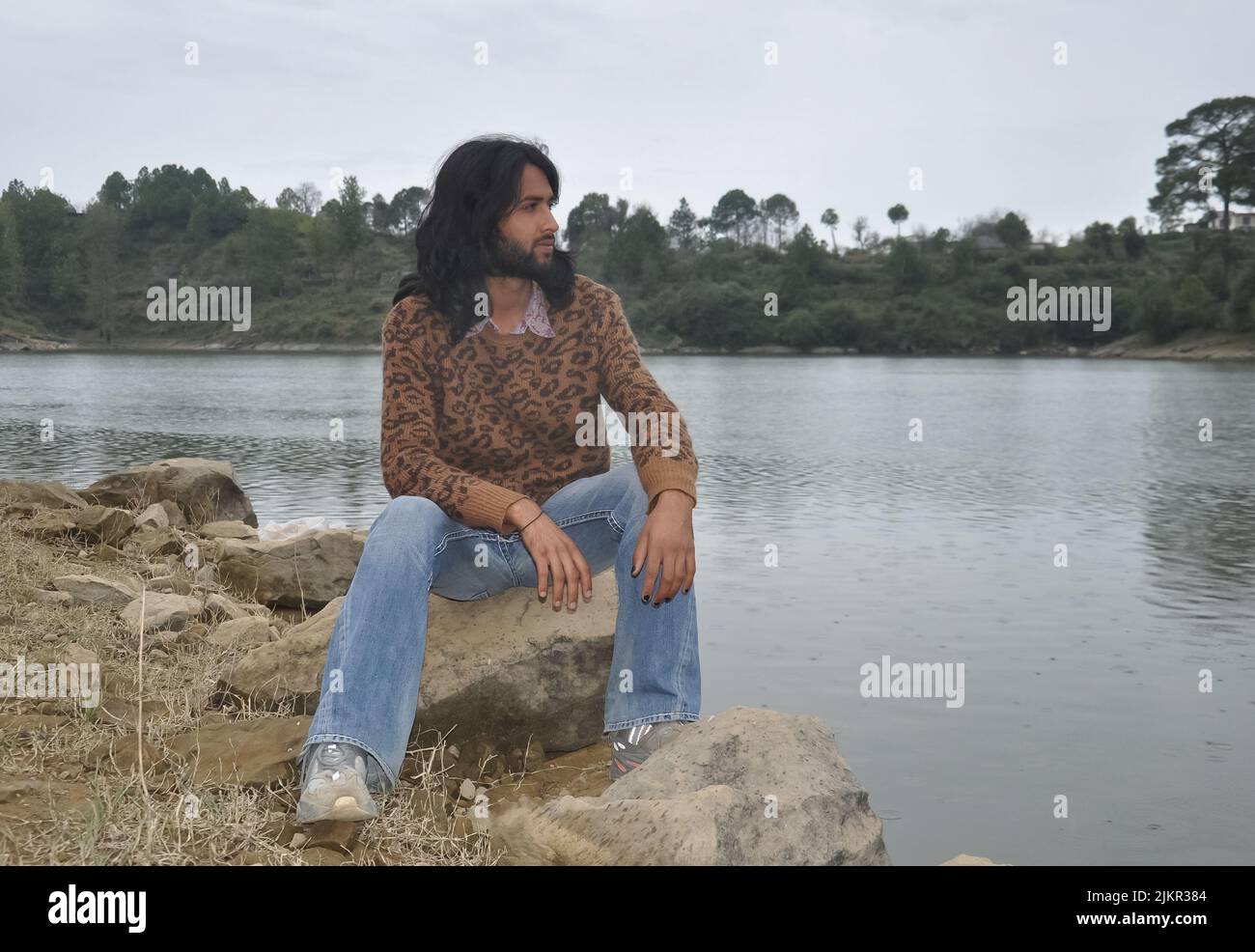 Un hombre joven de buena apariencia con pelo largo y barba mirando de lado mientras está sentado en piedra junto al lago Foto de stock