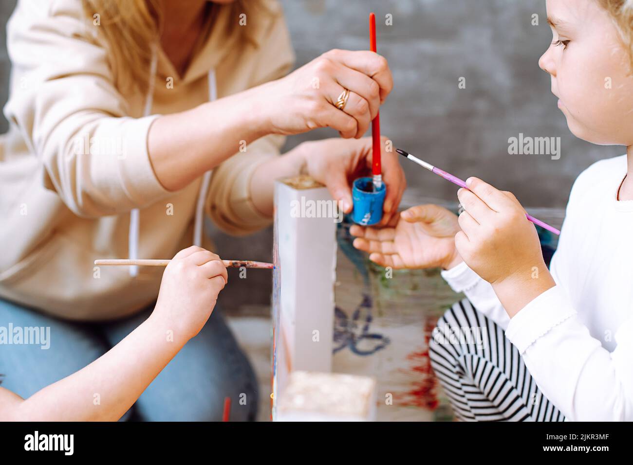 Foto recortada de la joven maestra que muestra cómo remover gouache azul con pincel rojo en un frasco de plástico a la niña. Foto de stock