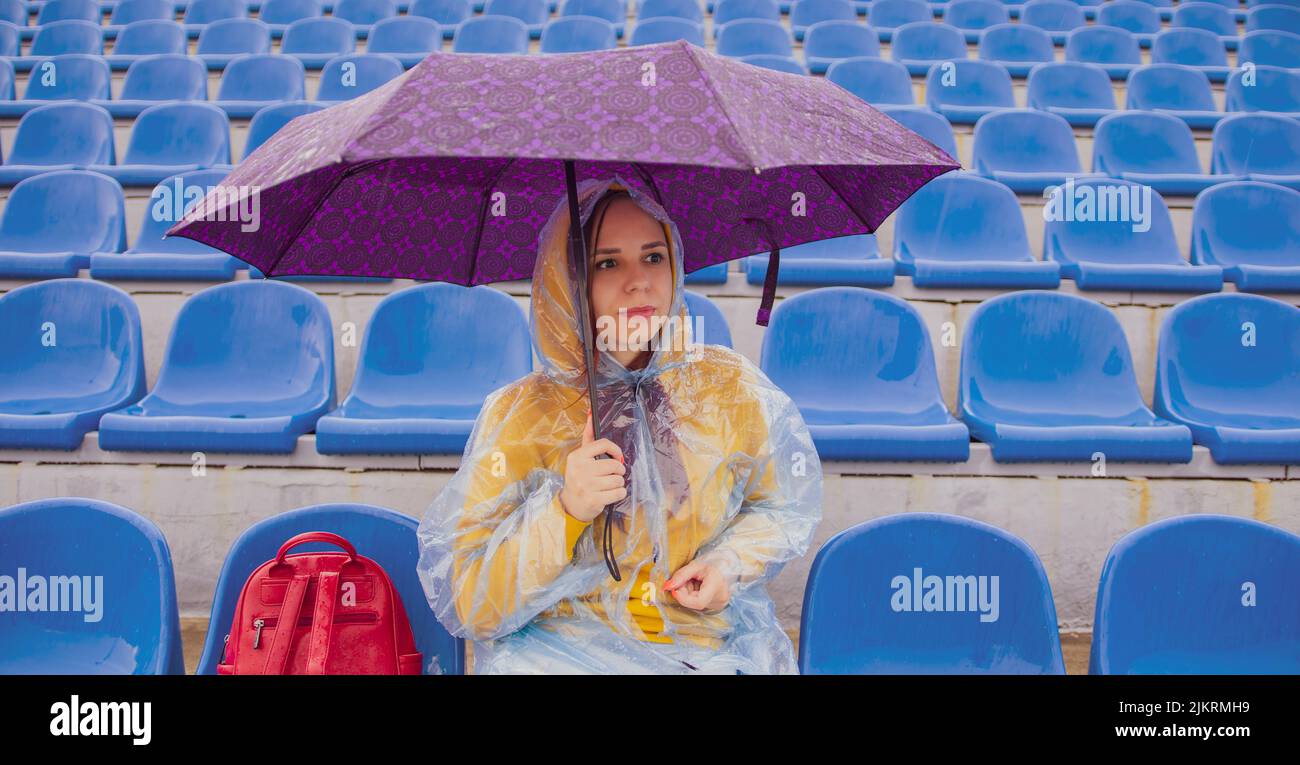 Joven con capucha y chubasquero de plástico mirando a un lado mientras se sienta en una tribuna con un paraguas en la mano durante el partido. Foto de stock