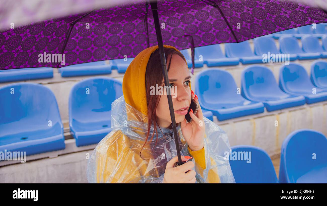 Una hermosa mujer con una sudadera con capucha y un impermeable de plástico, hablando por un teléfono móvil, sentada en el podio con un paraguas en la mano durante la lluvia. Foto de stock