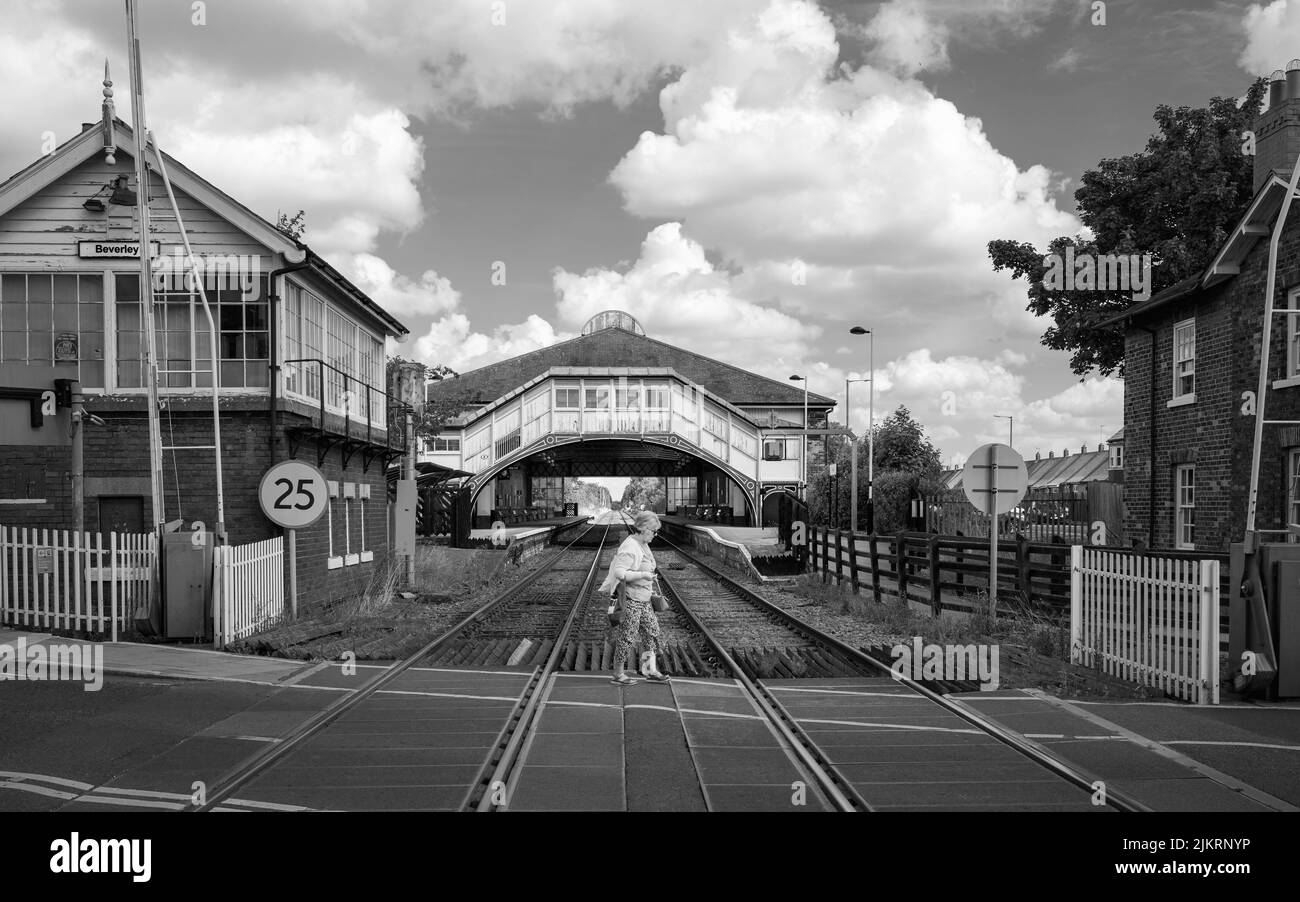 BEVERLEY, Reino Unido - 27 DE JULIO de 2022: Una mujer camina a través de las líneas de ferrocarril en las puertas batientes y la vista de la estación de ferrocarril en el fondo en Beverley, Reino Unido. Foto de stock