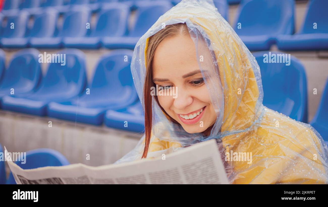 Una mujer hermosa en una sudadera con capucha y un impermeable de plástico, aprende las últimas noticias leyendo un periódico, sentado en el podio en un estadio vacío durante Foto de stock
