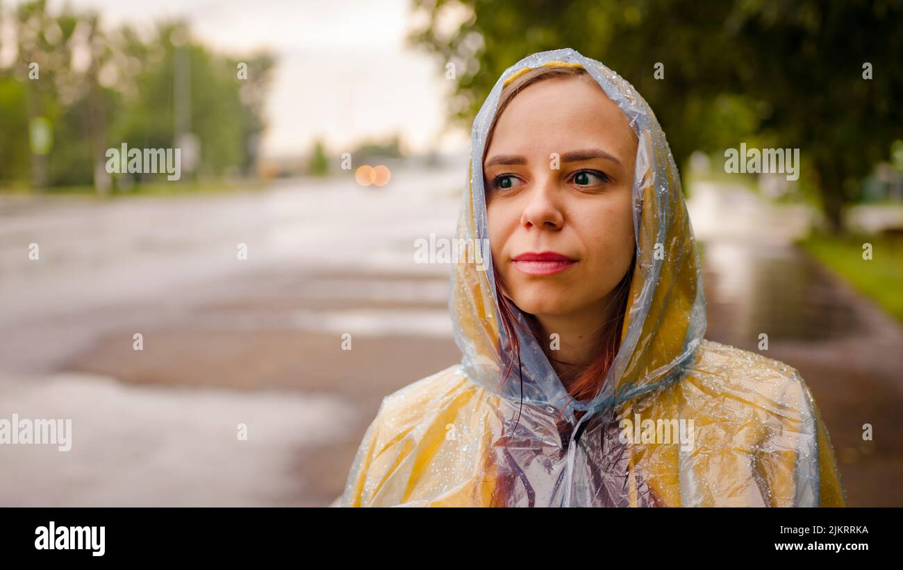 Mujer joven milenaria con capucha amarilla y impermeable transparente de pie en la carretera los días de lluvia en la ciudad. Foto de stock