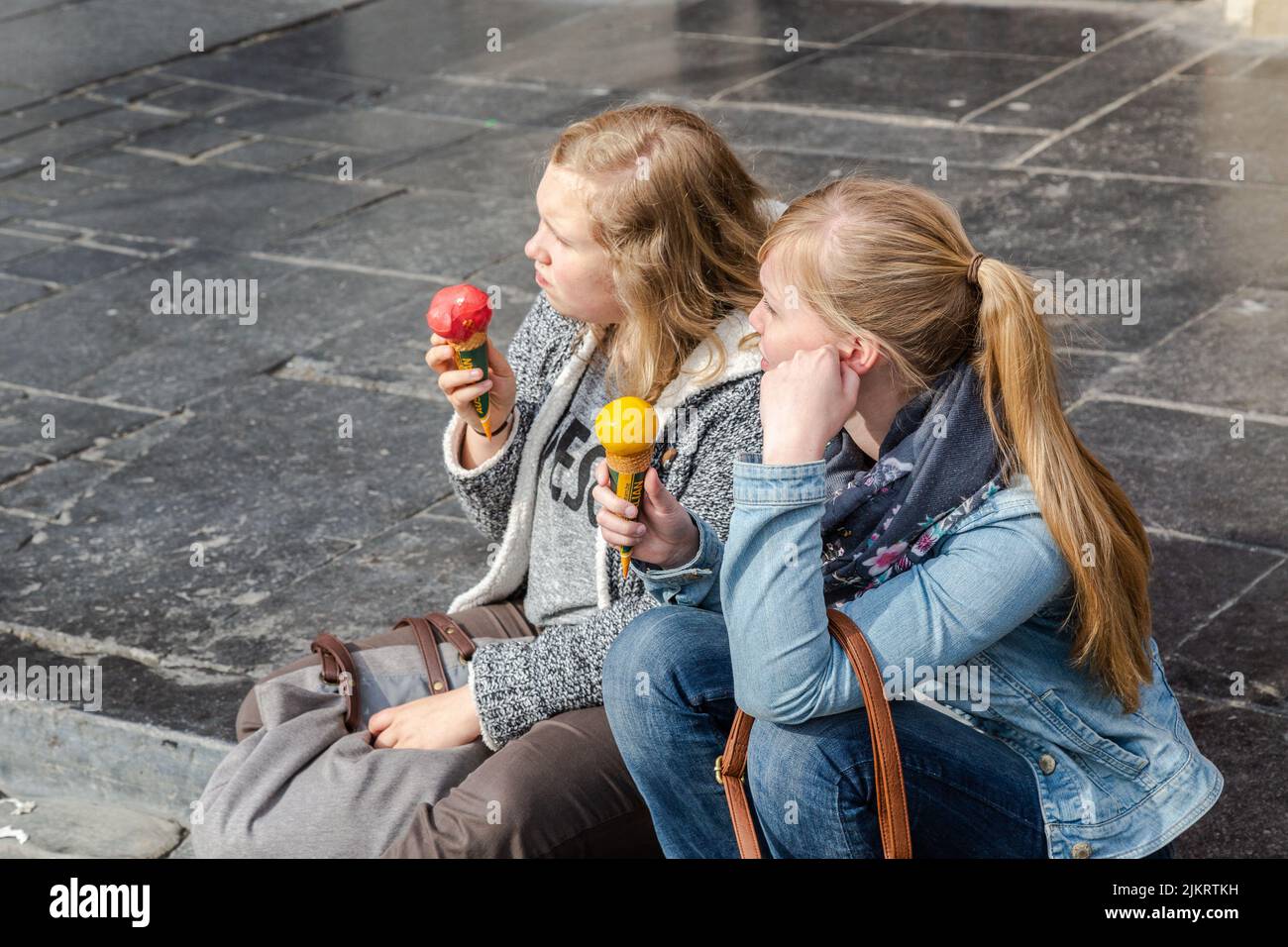 Dos mujeres jóvenes, portando un cono de helado, una cuchara roja, la otra amarilla, sentada en el borde de la acera. Foto de stock