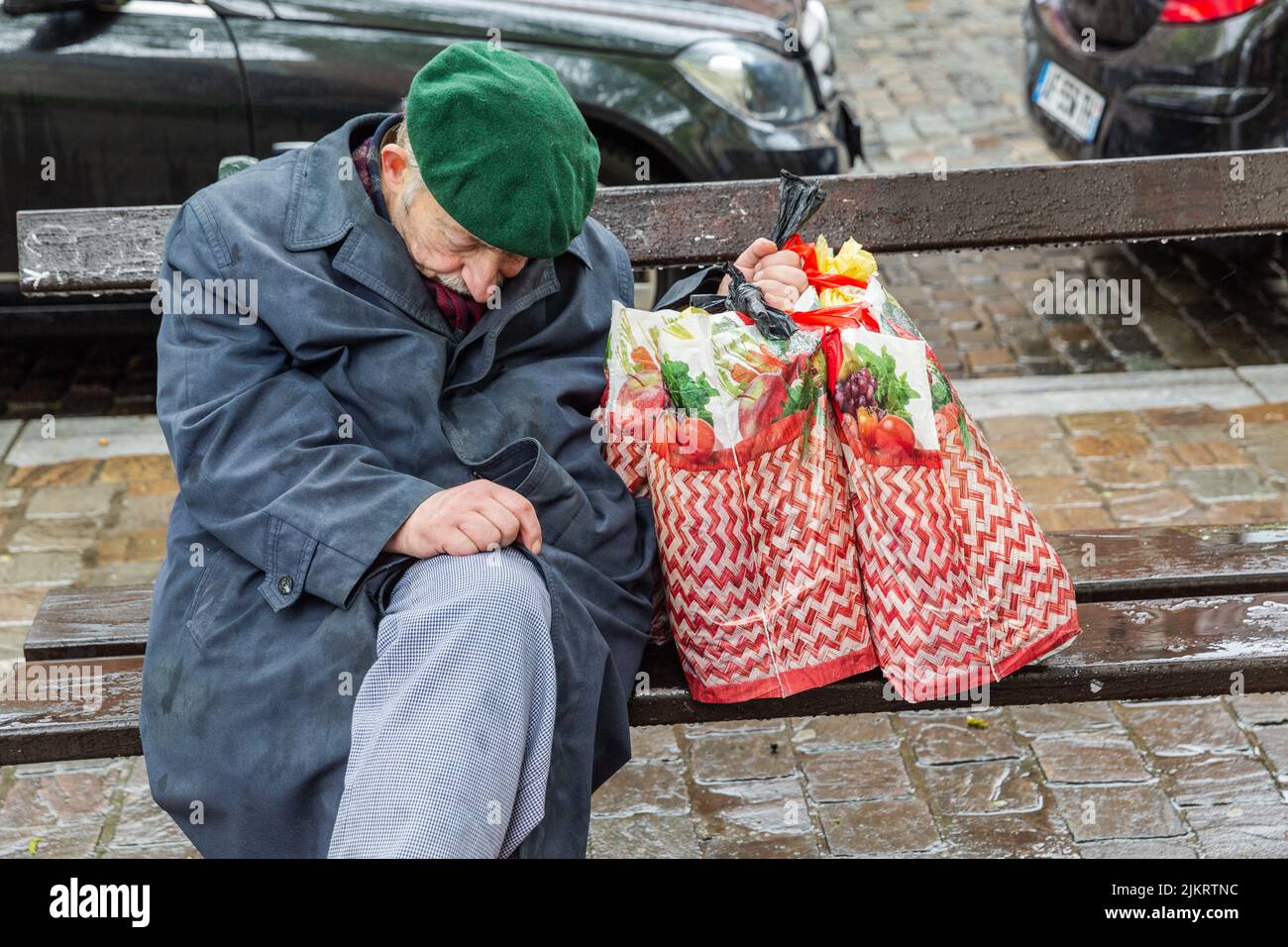 Anciano sentado en un banco público, somnoliento bajo la lluvia, sosteniendo sus dos bolsas de la compra en una mano. Foto de stock