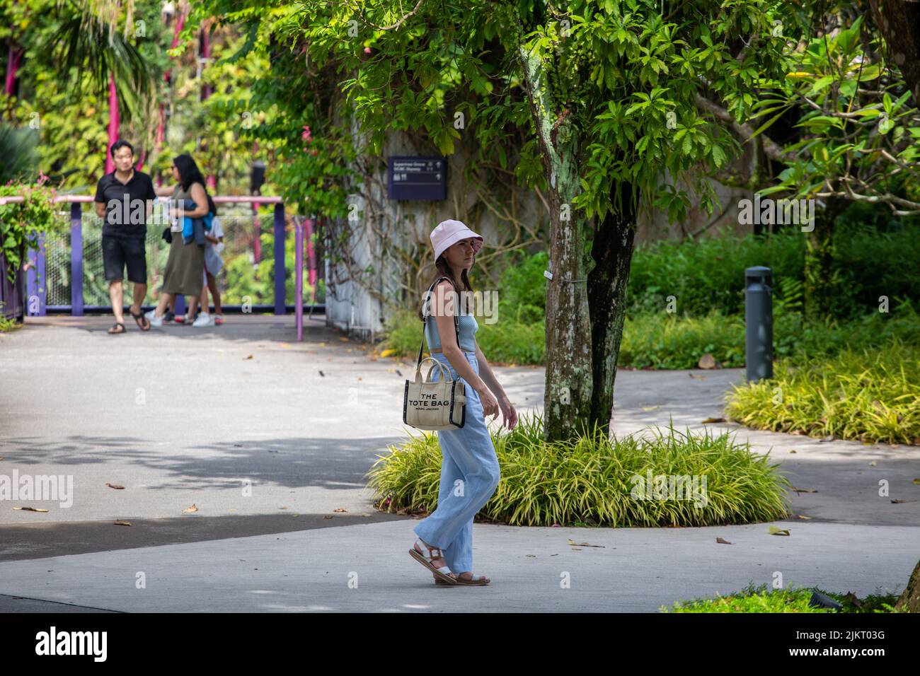 Un turista extranjero que lleva sombrero tipo cubo está recorriendo el Jardín junto a las instalaciones de la Bahía al aire libre pero a la sombra bajo el árbol. Singapur. 2022 Foto de stock
