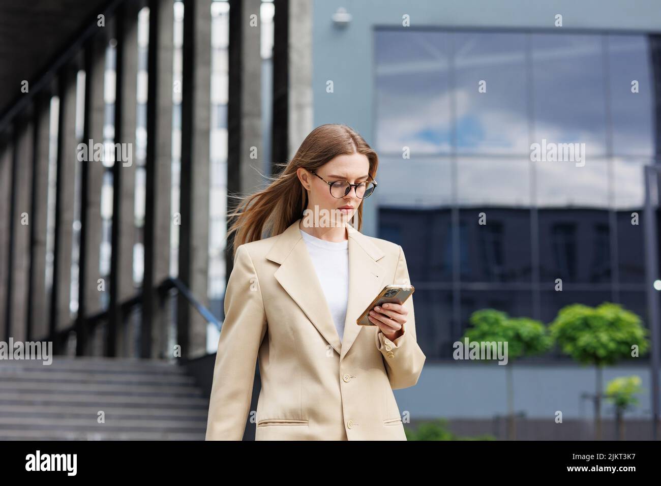Una joven rubia contra el telón de fondo de un centro de negocios, centro de oficinas. Mientras se desplaza, lee las noticias desde su smartphone. Foto de stock