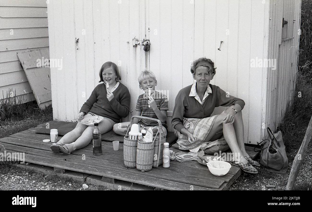 1960s, histórica, una madre con sus dos hijos pequeños sentados juntos en la plataforma exterior de madera de una cabaña de playa cerrada, habiendo encontrado un lugar nivelado para sentarse y su picnic de vacaciones, Inglaterra, Reino Unido. Foto de stock