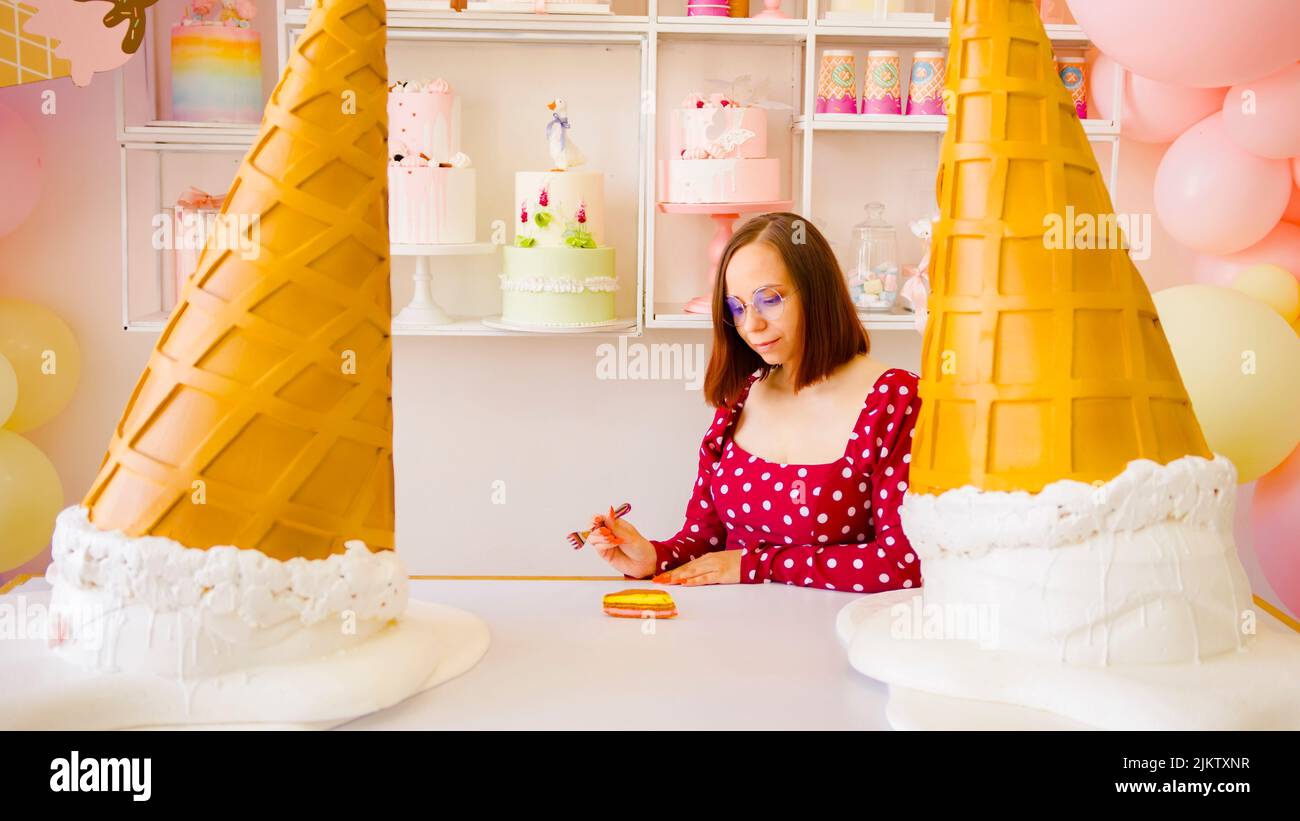 Mujer joven en vasos comiendo pastel con tenedor, de pie en vívida confitería. Mujer feliz en vestido rojo masticando comida dulce, disfrutando Foto de stock