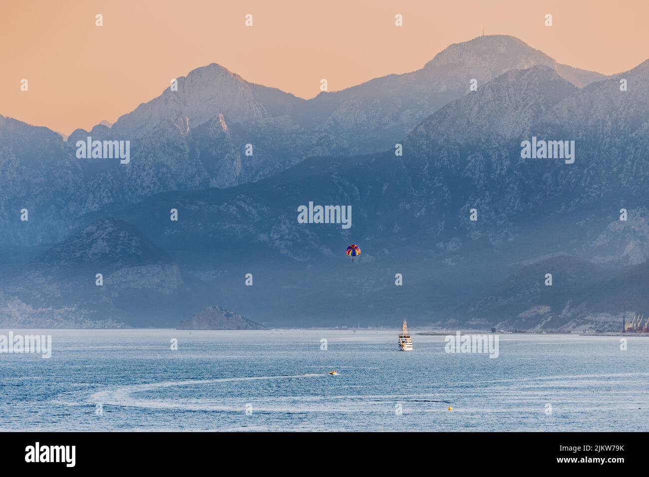 Vista de zoom distante de un barco turístico de crucero y paracaídas de deportes acuáticos con majestuosas montañas Tauro en la región de Antalya en Turquía Foto de stock