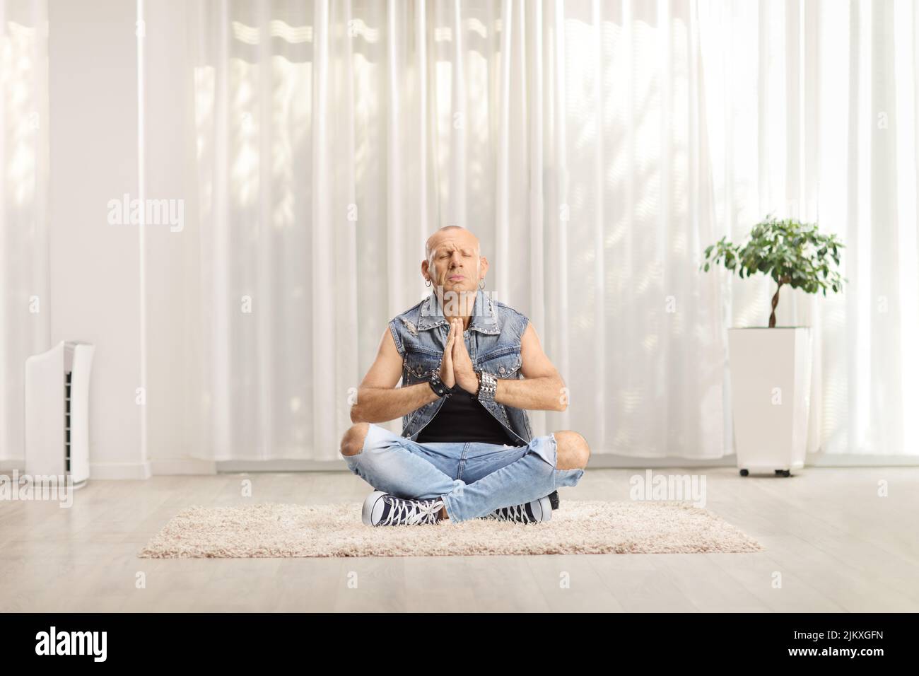Hombre calvo con jeans sentado con piernas cruzadas y practicando yoga en casa sobre una alfombra Foto de stock