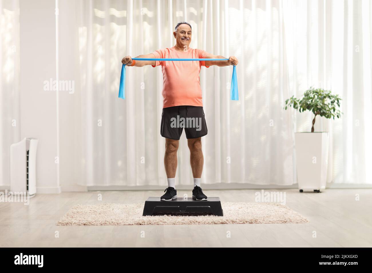 Hombre maduro en sportswear haciendo ejercicio con una correa elástica en una plataforma aeróbica paso dentro de una habitación Foto de stock