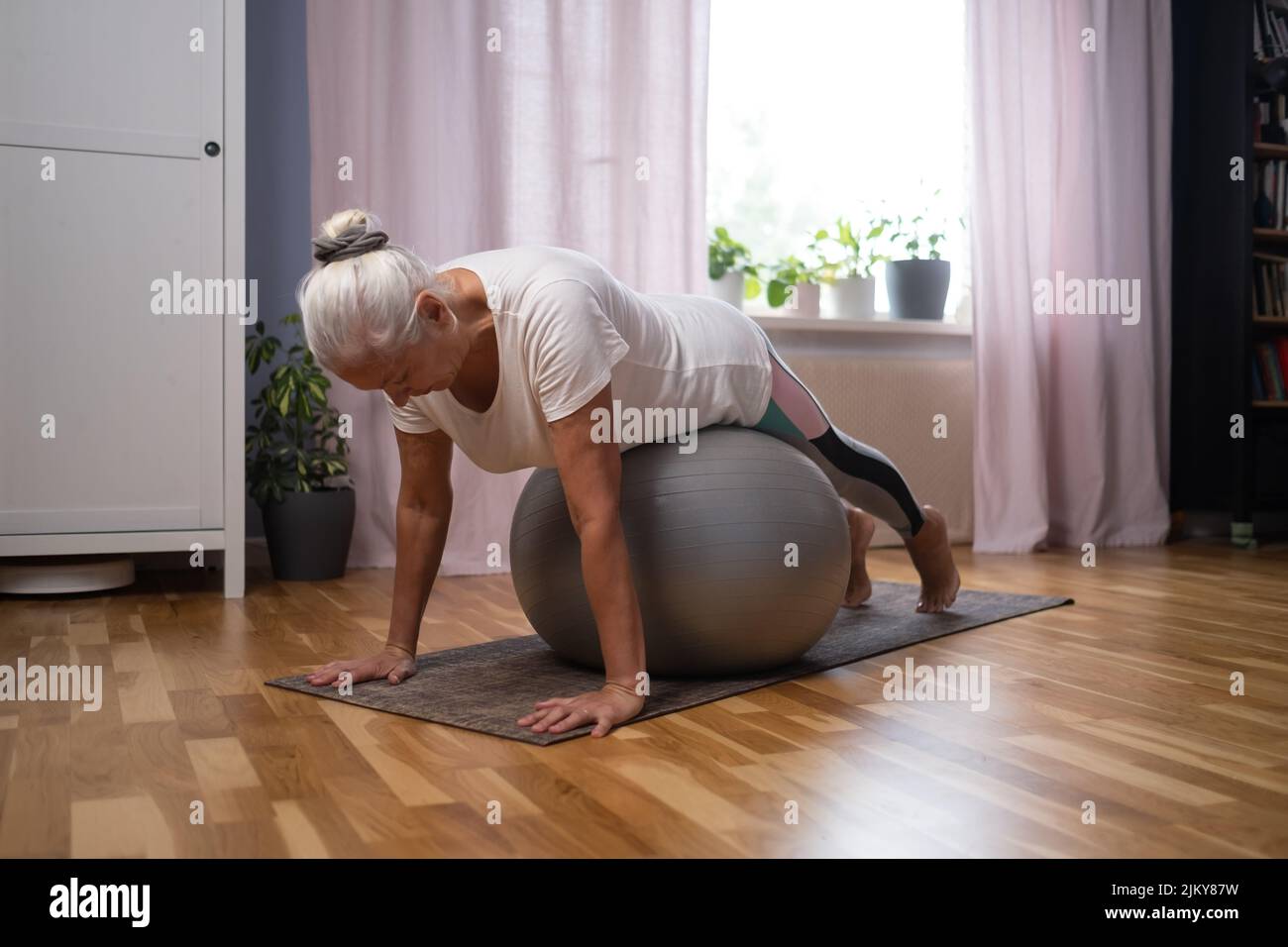 Entrenamiento en casa. Mujer mayor deportiva haciendo una tabla de yoga con una pelota haciendo ejercicio en la sala de estar Foto de stock