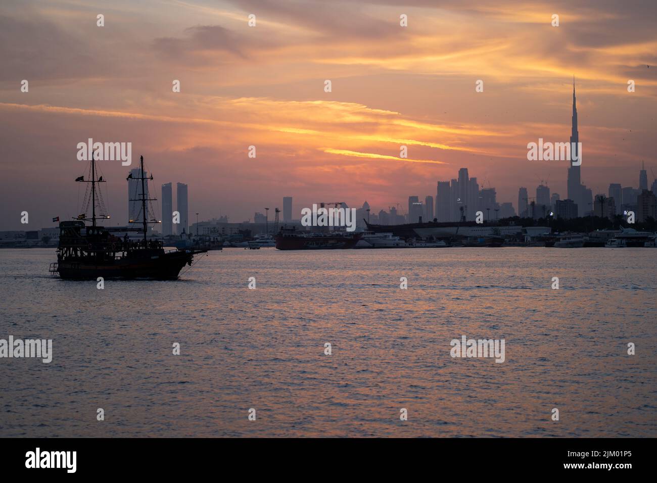 Una vista impresionante de un barco en Festival City Lake con el paisaje urbano de Dubái al atardecer como fondo Foto de stock