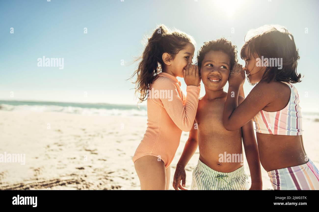 Dos juguetonas chicas susurrando en los oídos de su amiga en la playa. Grupo de niños adorables divirtiéndose y disfrutando de sus vacaciones de verano Foto de stock