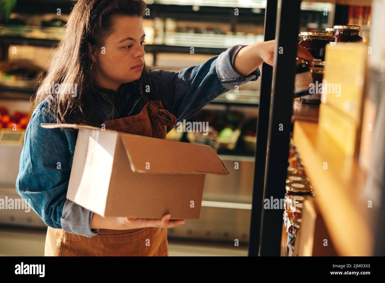 Empleado del supermercado con síndrome de Down reabasteciendo los productos alimenticios en los estantes de la tienda. Mujer empoderada con una discapacidad intelectual que trabaja como a. Foto de stock