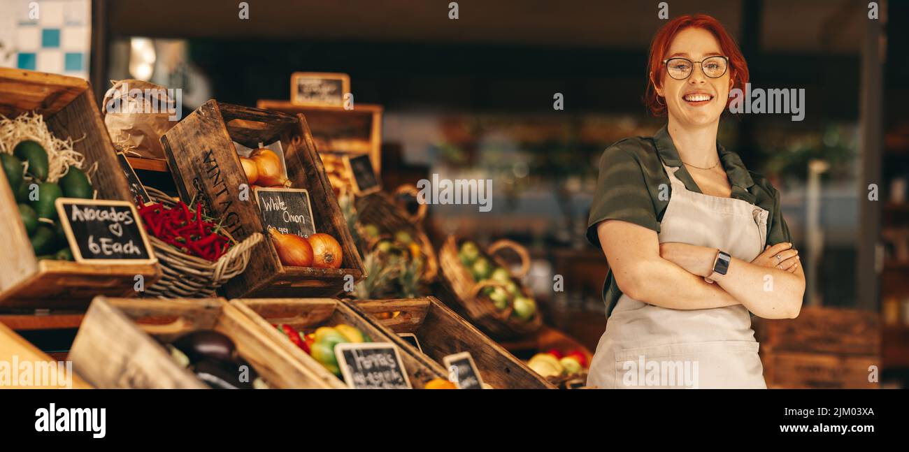 Feliz propietario del supermercado sonriendo a la cámara mientras se encuentra en la sección de productos frescos de su tienda de comestibles. Mujer joven exitosa corriendo un pequeño Foto de stock