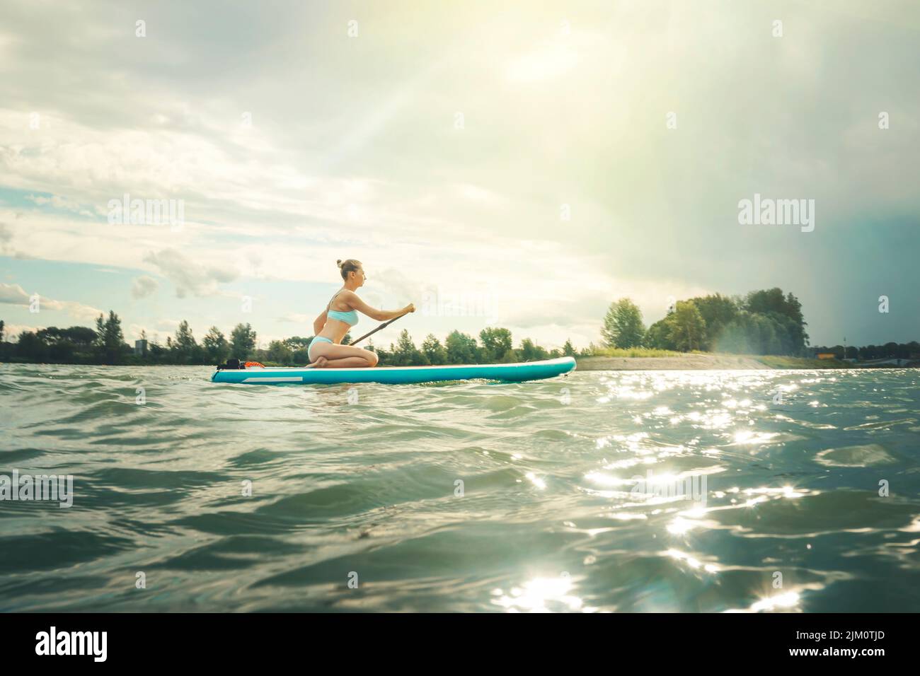 Feliz hermosa mujer atlética en un traje de baño azul con una paleta en sus manos en una tabla de surf en un día soleado en el agua del mar azul. Concepto de A. Foto de stock