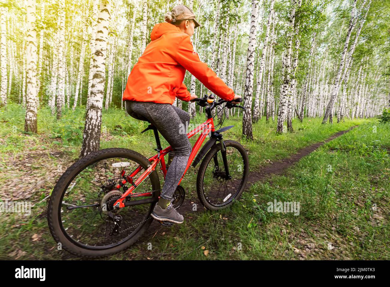 Mujer joven deportiva con chaqueta naranja brillante y vaqueros monta una bicicleta por el bosque en un día de verano. Vista posterior Foto de stock