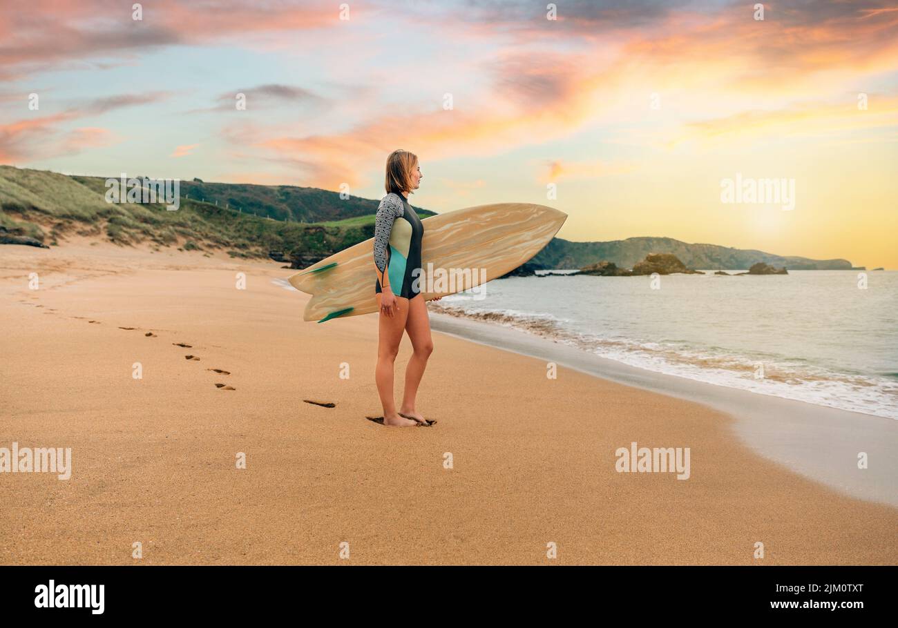 Mujer surfista con traje de neopreno llevando tabla de surf mirando al mar en la playa Foto de stock