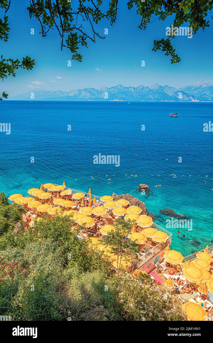 23 de junio de 2022, Antalya, Turquía: Playa estrecha y acogedora escondida entre acantilados y rocas con sombrillas de color amarillo brillante y tumbonas y un pintoresco azul refrescante Foto de stock