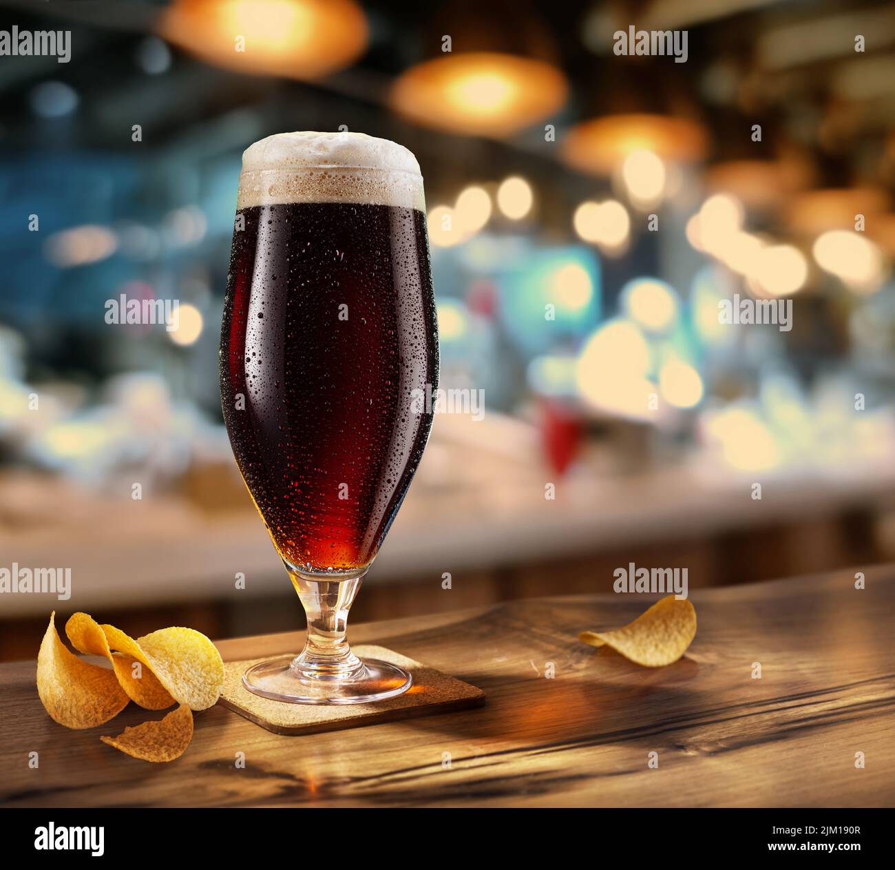 Vaso frío de cerveza oscura sobre la mesa de madera. Interior de pub borroso al fondo. Foto de stock