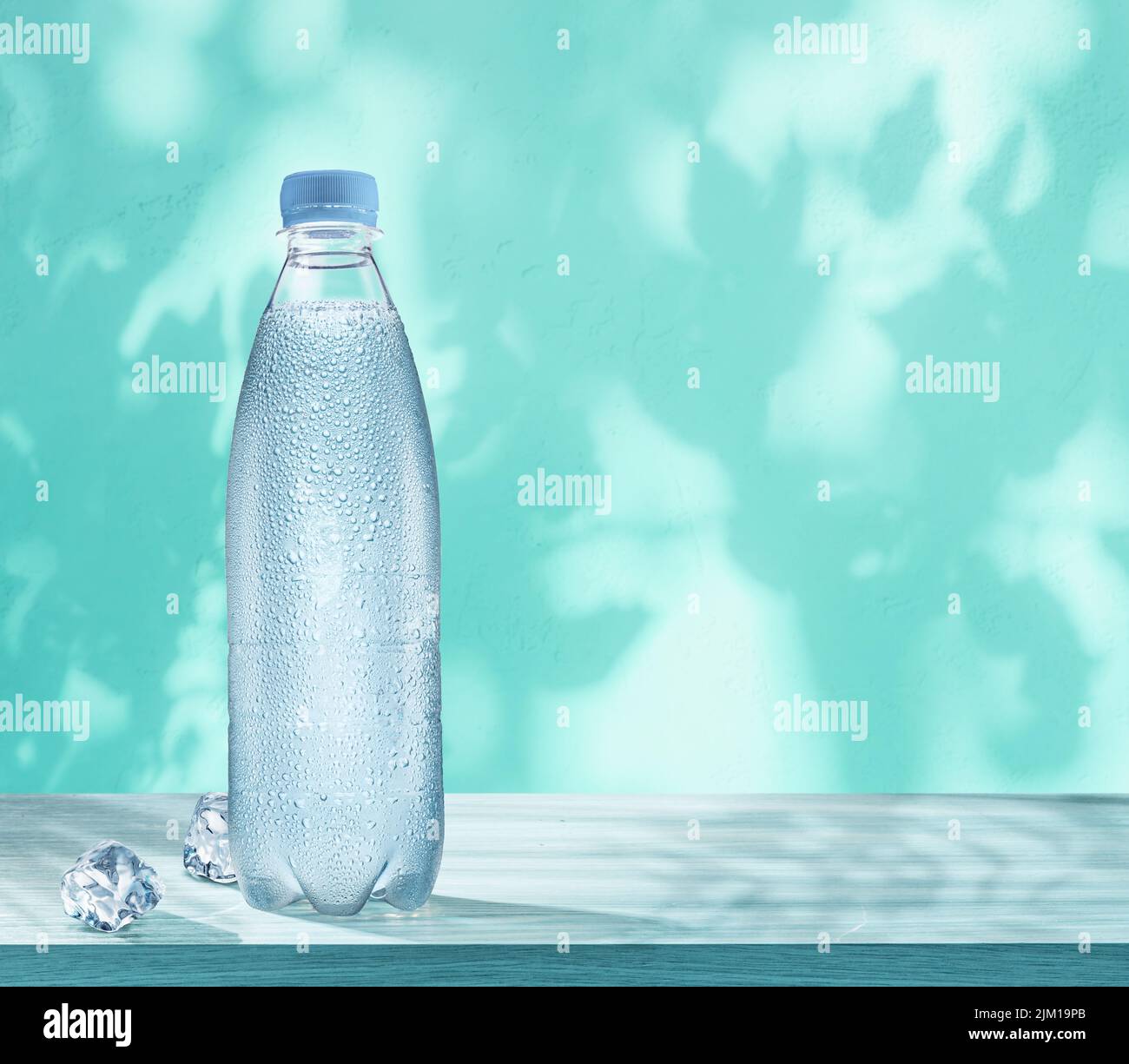 Botella de plástico de agua fría con gotas de condensación y dos cubitos de hielo sobre la mesa. Pared con sombras de hojas en el fondo. Foto de stock