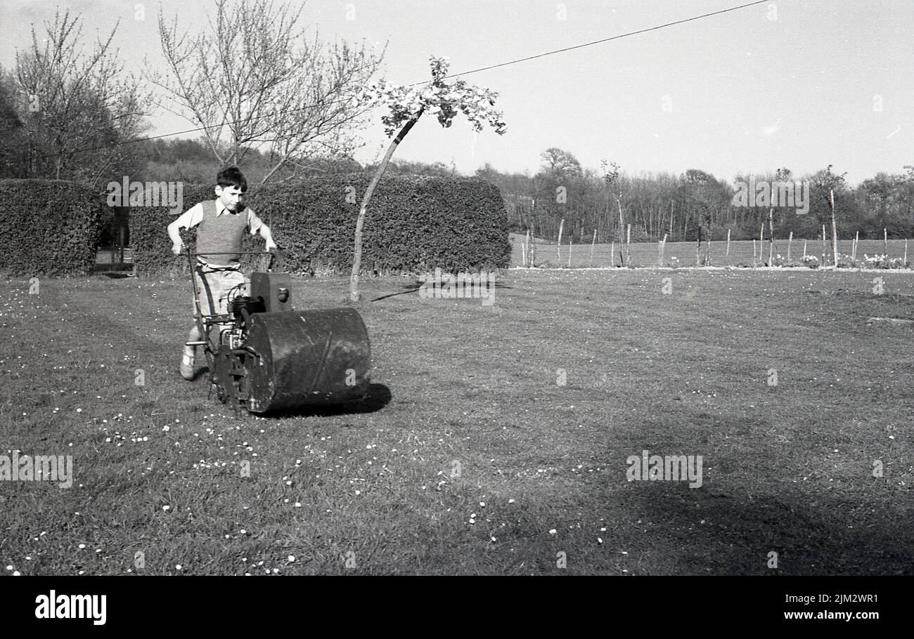 1960s, histórico, fuera en un jardín grande, un niño joven que corta un césped, empujando un viejo cortacésped diesel grande, Inglaterra, Reino Unido. Foto de stock
