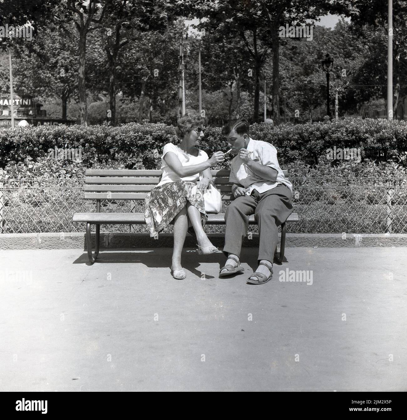 1950s, histórico, un día soleado y una señora sentada en un banco al lado de un hombre, encendiendo su cigarrillo, usando su propio cigarrillo encendido, Inglaterra, Reino Unido. Foto de stock