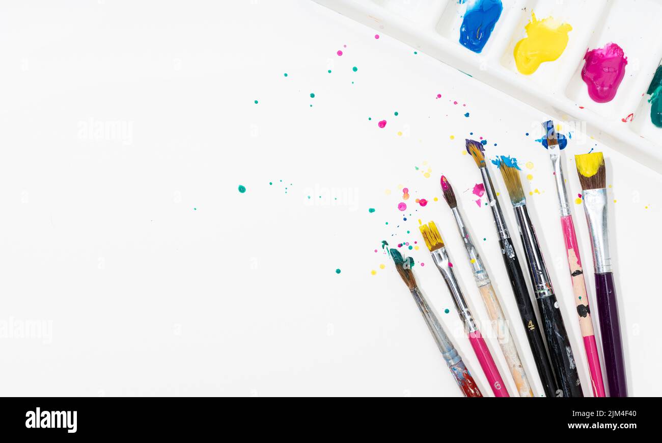 Arte Pinceles de pintura y paleta con salpicadura de pintura de colores sobre papel de lienzo blanco. Espacio de copia Foto de stock