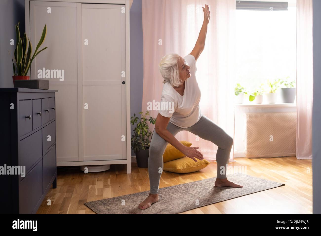 Feliz señora mayor haciendo yoga asana en la sala de estar. Foto de stock