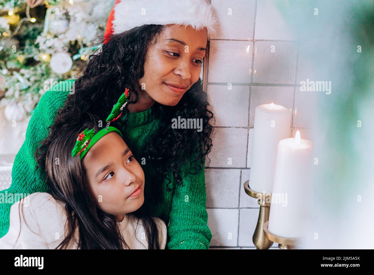 Mujer afroamericana en sombrero de Santa y morena hija de pelo largo posan contra decoraciones navideñas y velas sonriendo alegremente Foto de stock
