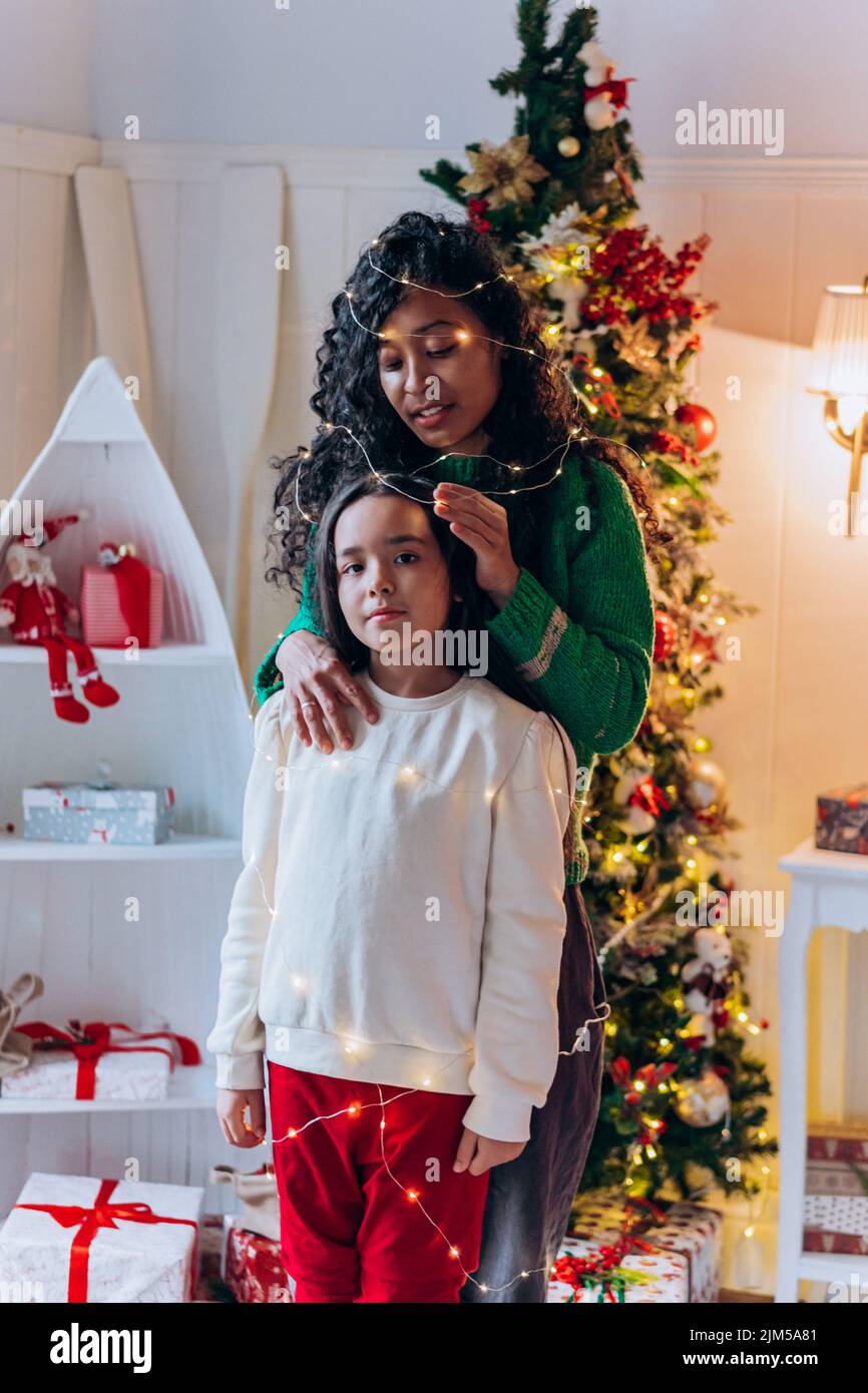 Una niña morena y una madre afroamericana de pelo rizado posan con guirnalda iluminada en la cabeza y sonríen alegremente contra el árbol de Navidad Foto de stock
