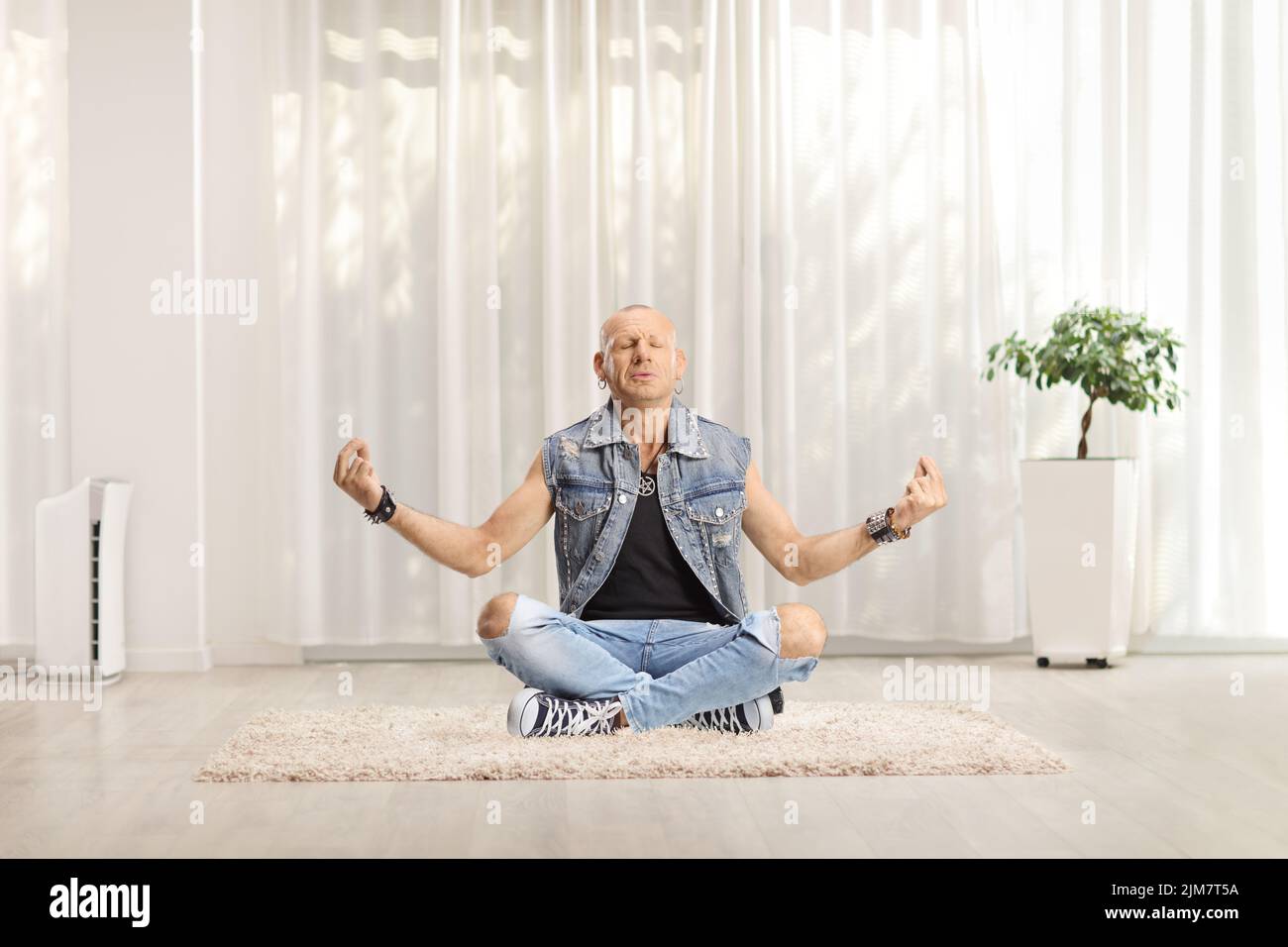 Hombre con vaqueros sentado en una alfombra practicando meditación en una sala de estar en casa Foto de stock